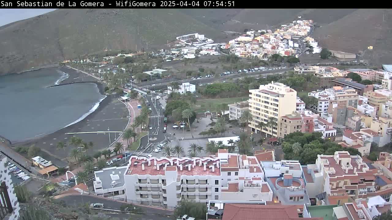 An aerial view shows a town situated on a volcanic hillside overlooking a dark-sand beach and calm ocean under an overcast sky.