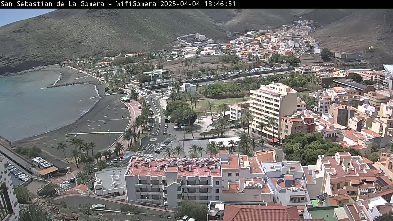 A coastal town with a black sand beach and numerous buildings stretches along a hillside under a mostly clear sky.