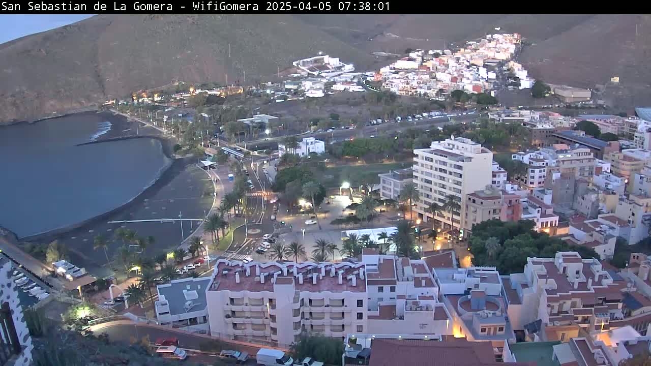 An aerial view of a coastal town at dusk, showing a dark-colored beach, buildings, palm trees, and a calm body of water.