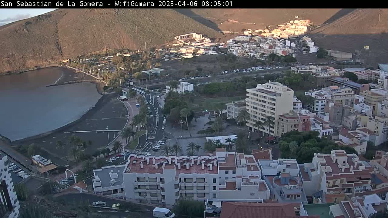 An aerial view shows a coastal town with a dark-sand beach and calm water, under mostly sunny skies with some shadows on the hills in the background.
