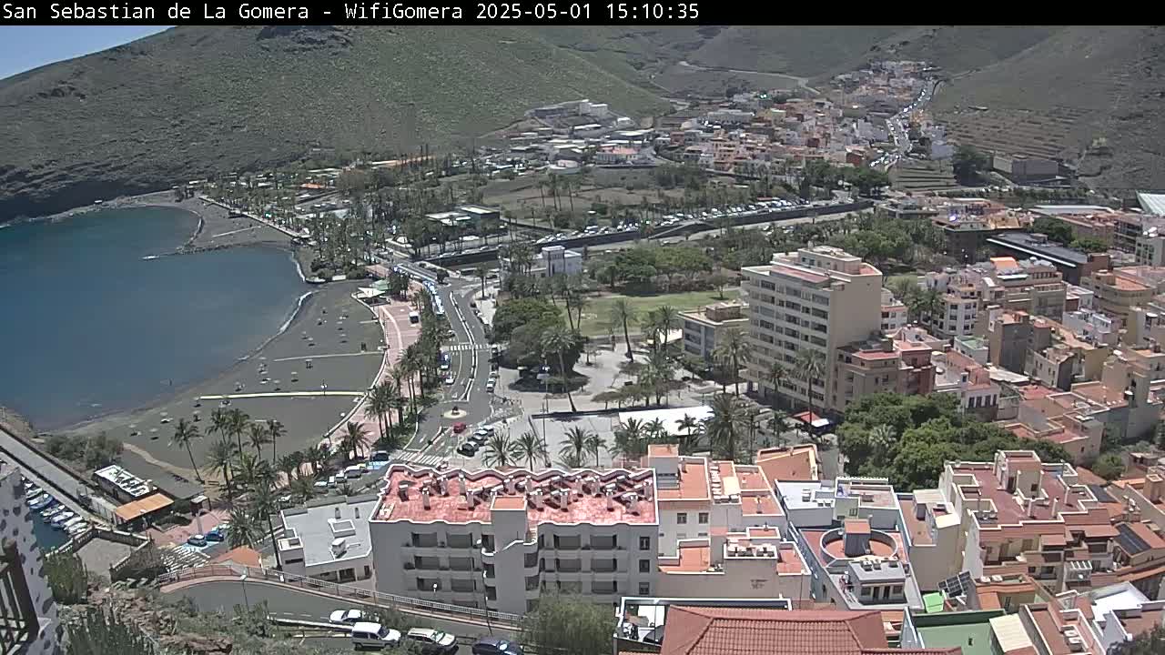 A coastal town is seen from above on a sunny day, featuring a dark-sand beach, calm bay, palm trees, buildings, and roads.