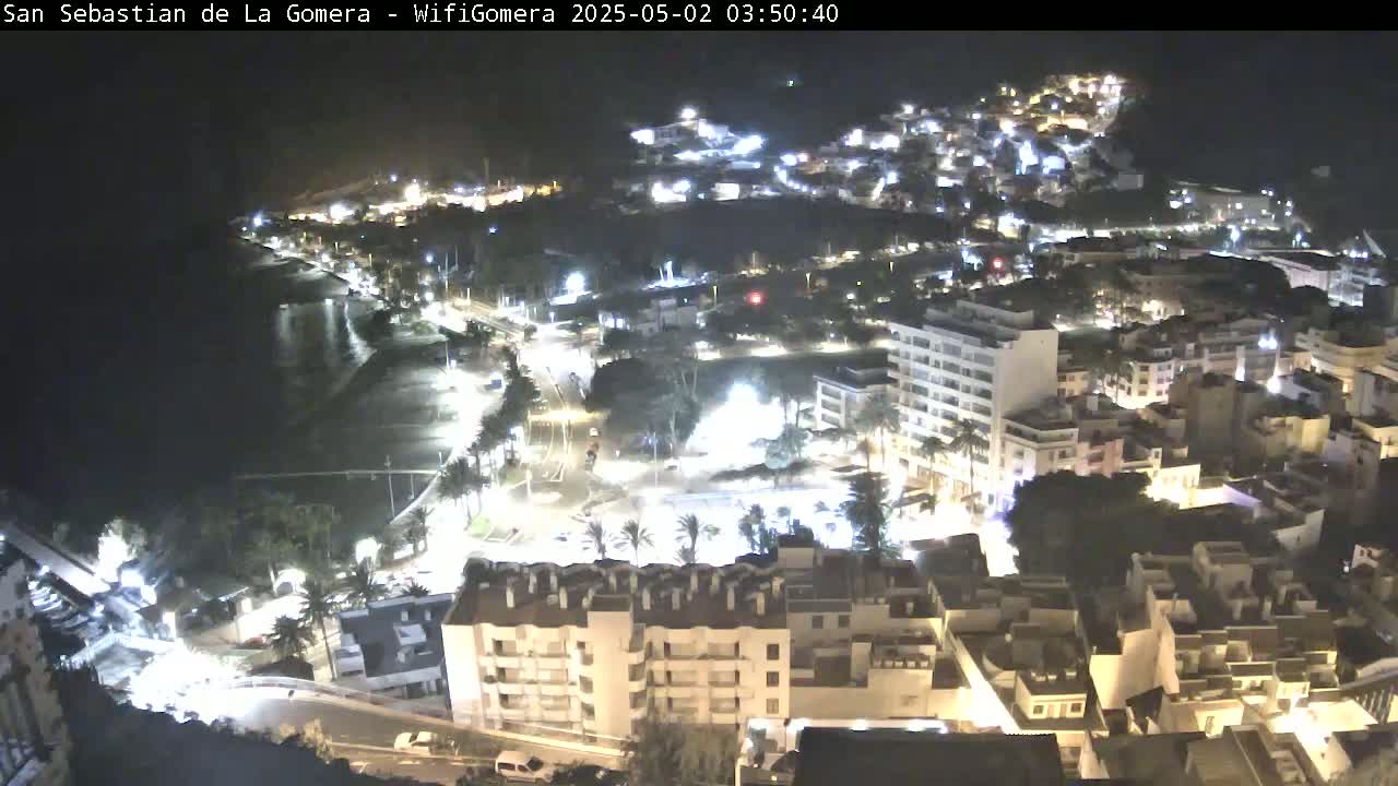 A nighttime aerial view of a coastal town with many buildings lit up, and a dark ocean visible in the background.