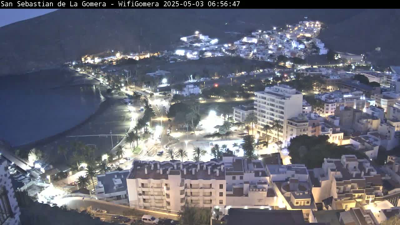 A coastal town at night, illuminated with streetlights, is nestled beside a dark body of water under a twilight sky.
