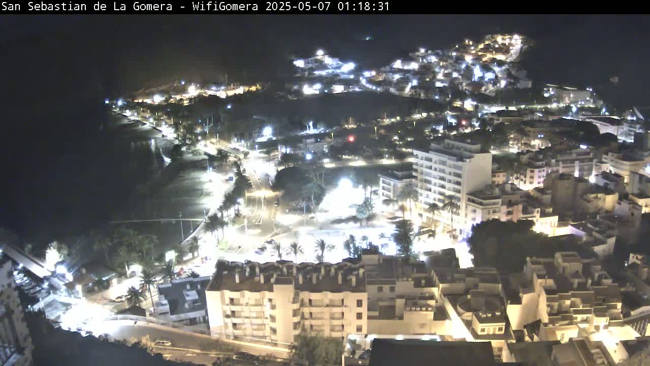 A nighttime aerial view shows a coastal town with brightly lit buildings, streets, and a beach, under a clear, dark sky.