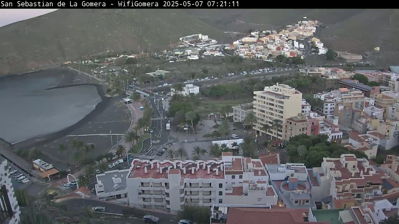 An aerial view shows a town situated on a hillside overlooking a dark-sand beach and calm ocean under a twilight sky.