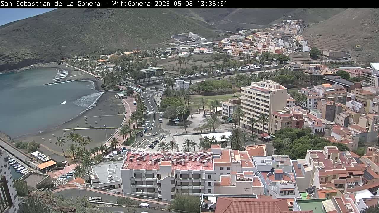 An aerial view shows a coastal town nestled against a mountainside on a sunny day, with a dark-sand beach and calm ocean visible below.