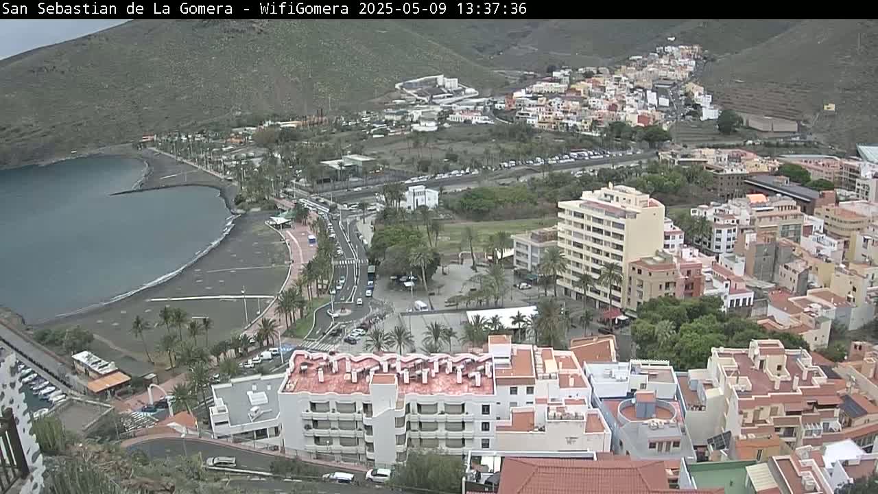 An aerial view shows a coastal town on a partly cloudy day, with a dark-sand beach bordering a calm bay, and numerous buildings extending up a hillside.