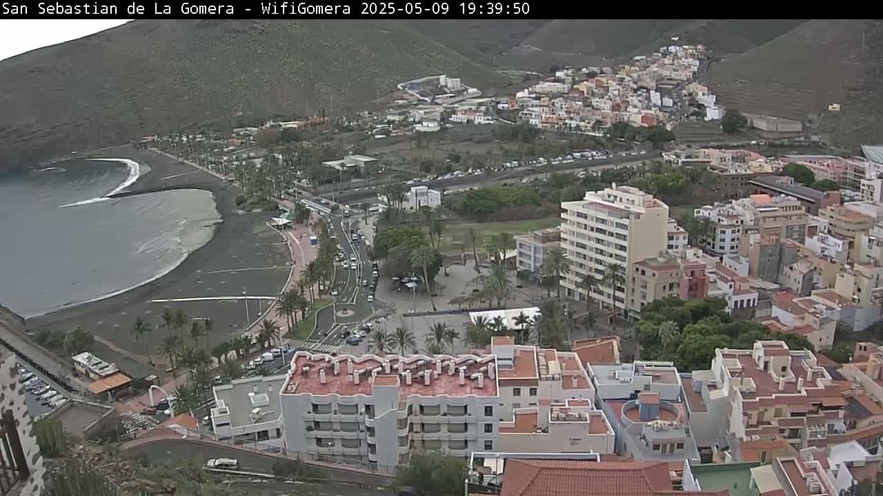 An overcast view of a town situated on a coast with a dark-sand beach and calm ocean waves.