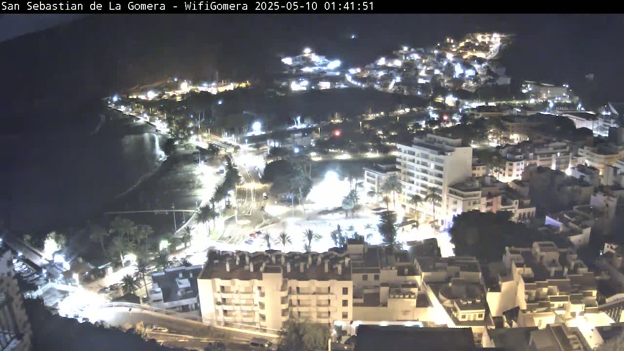 A nighttime aerial view shows a coastal town with buildings illuminated against a dark ocean.