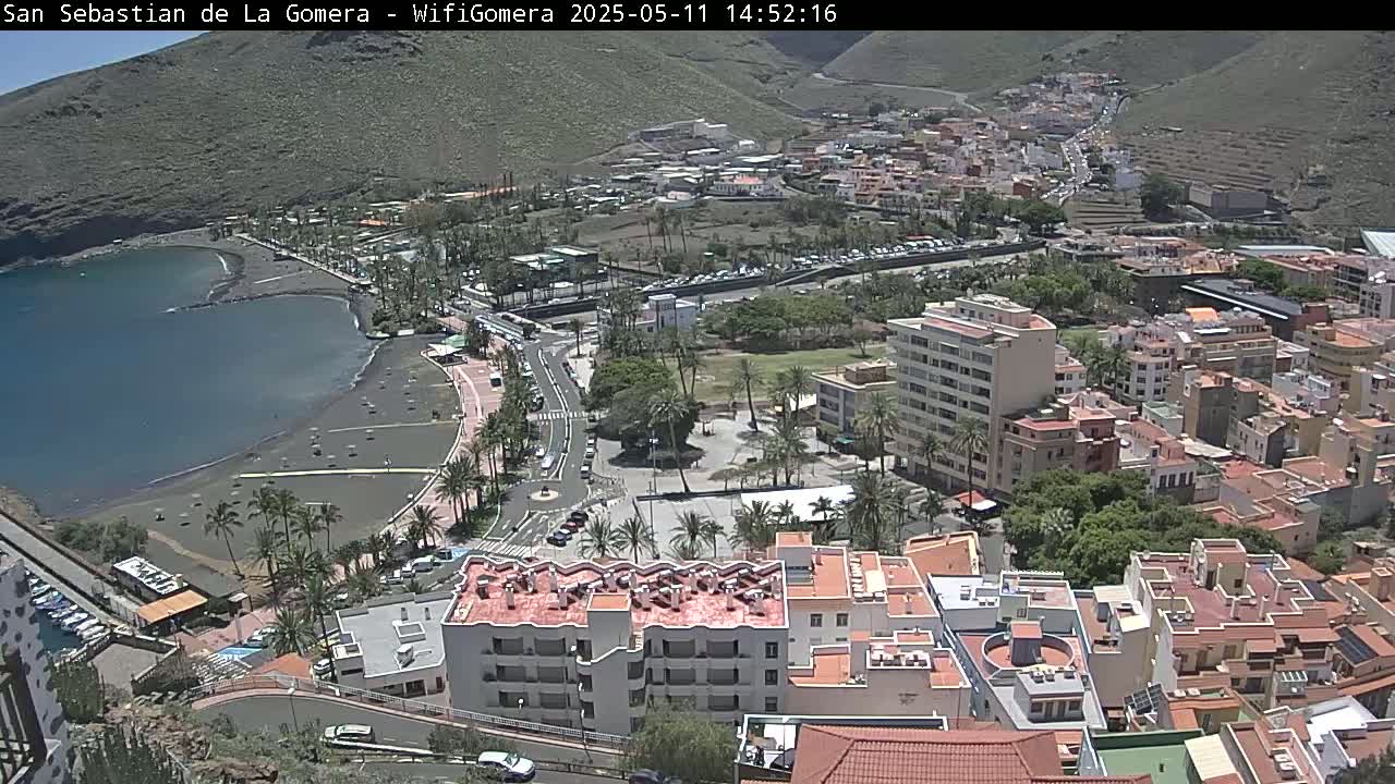 An aerial view shows a coastal town on a sunny day, featuring a dark-sand beach bordering a calm bay, and buildings stretching up a hillside.