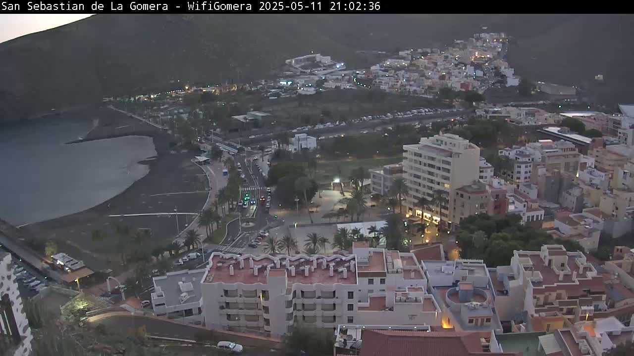 An aerial view at dusk shows a town with buildings clustered around a dark-sand beach and calm water, situated against a dark mountainside.