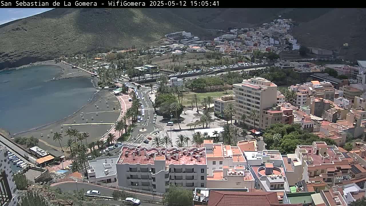 A coastal town with a black sand beach is seen from above on a sunny day, surrounded by buildings and palm trees against a backdrop of hills.