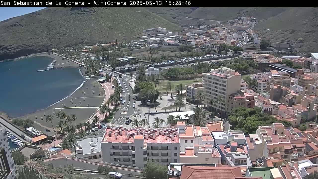A coastal town with a dark-sand beach and numerous buildings is viewed from above on a sunny day.