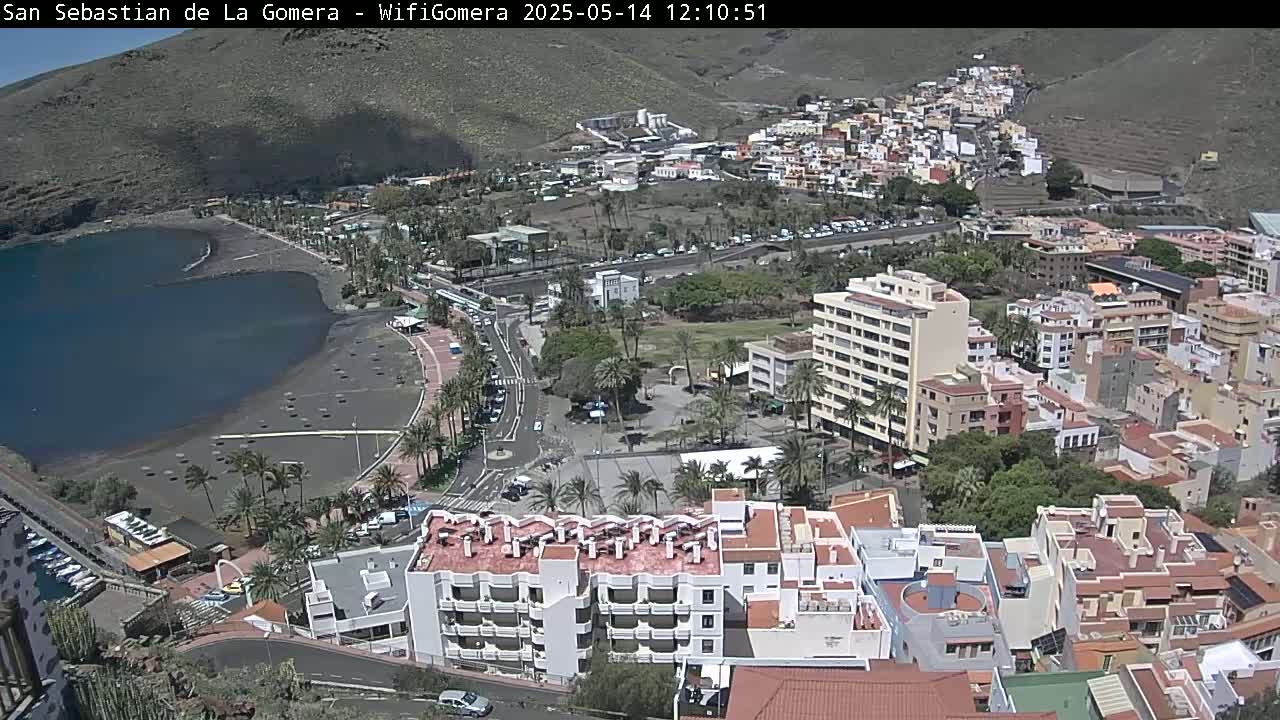 A coastal town with a dark-sand beach and calm bay, surrounded by buildings under a sunny sky.