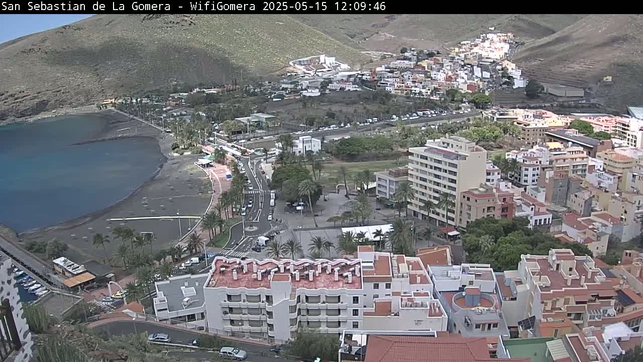 A coastal town with a black sand beach, buildings clustered together on a hillside, and a partly cloudy sky.