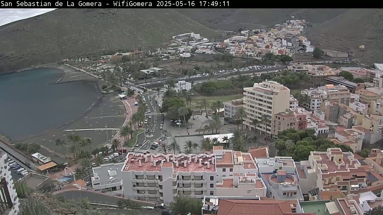 An aerial view of a coastal town on a partly cloudy day, showing a dark-sand beach, buildings clustered on a hillside, and palm trees lining a promenade.
