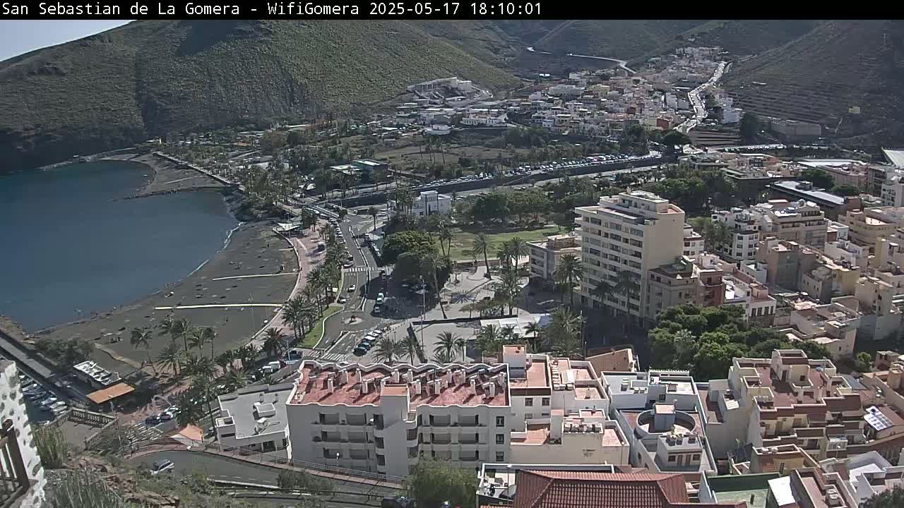 A coastal town nestled against a mountainside, featuring a dark-sand beach, calm waters, and buildings under a sunny sky.