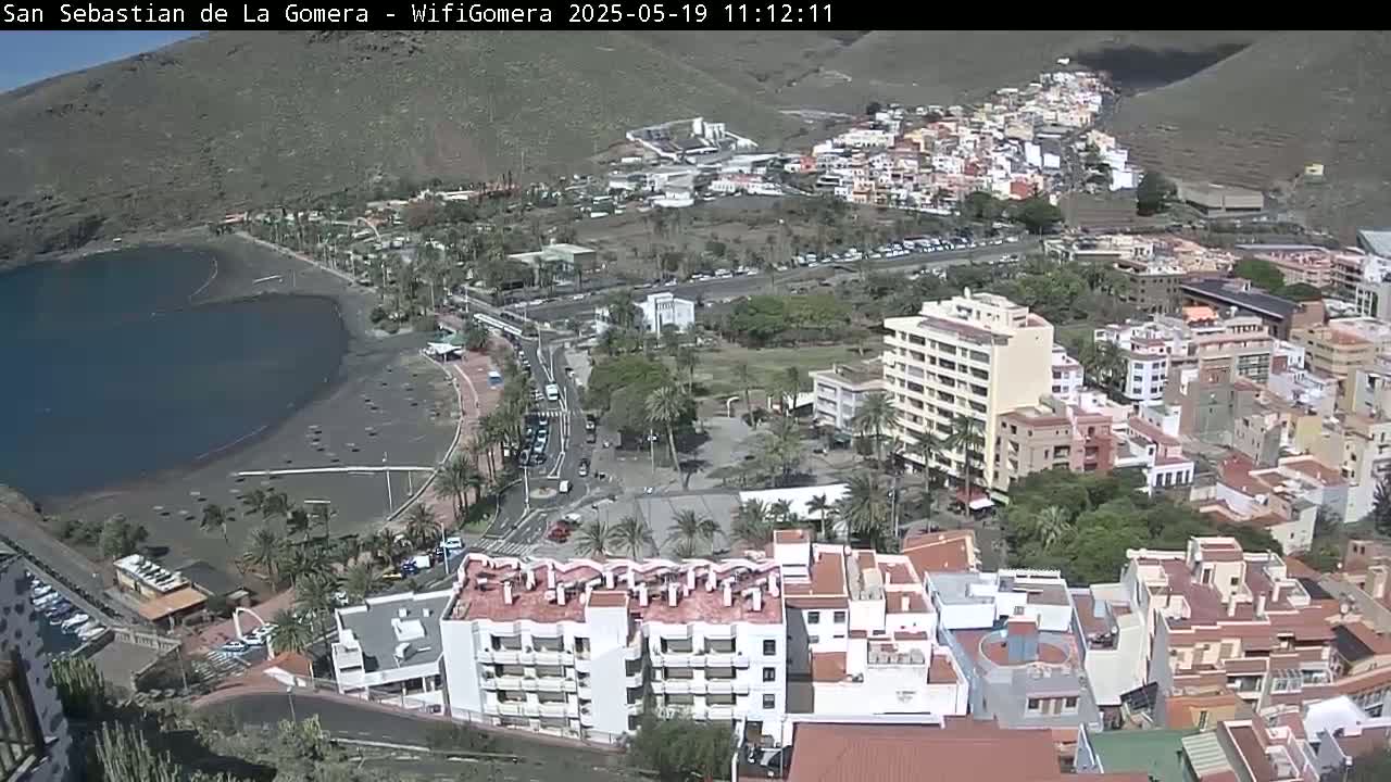 A coastal town with dark sand beach and calm water is nestled below a hillside, with numerous buildings and palm trees visible on a sunny day.