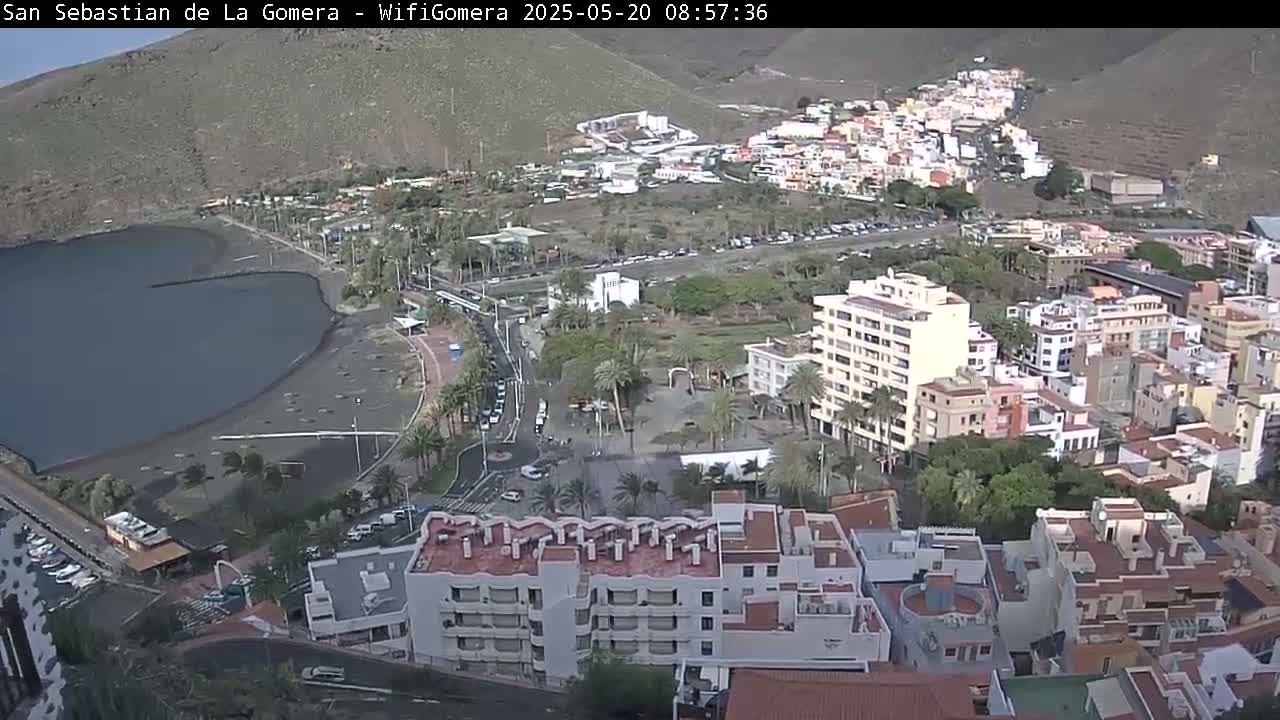A coastal town with dark-sand beaches and numerous buildings is seen from an elevated viewpoint under a mostly clear sky.