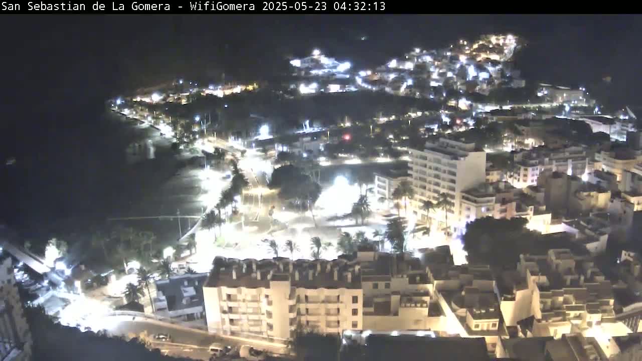 A nighttime aerial view of a coastal town shows a beach, buildings illuminated by lights, and calm waters.