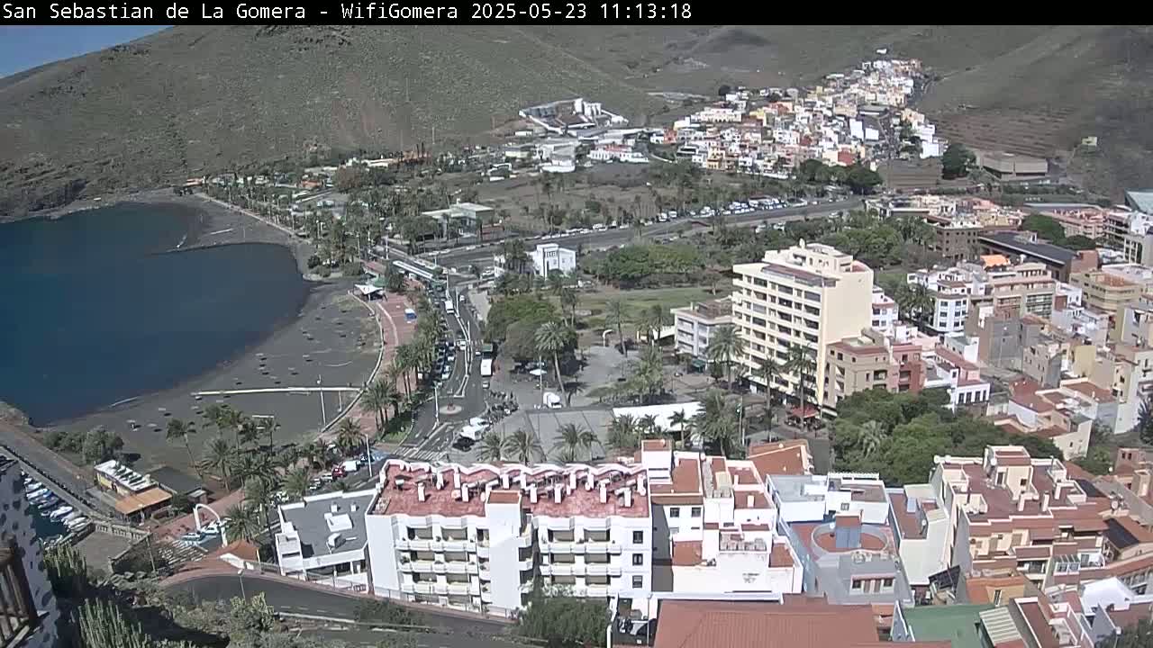 A coastal town with a dark-sand beach and calm ocean, nestled between hillsides under sunny skies, is viewed from an elevated perspective.