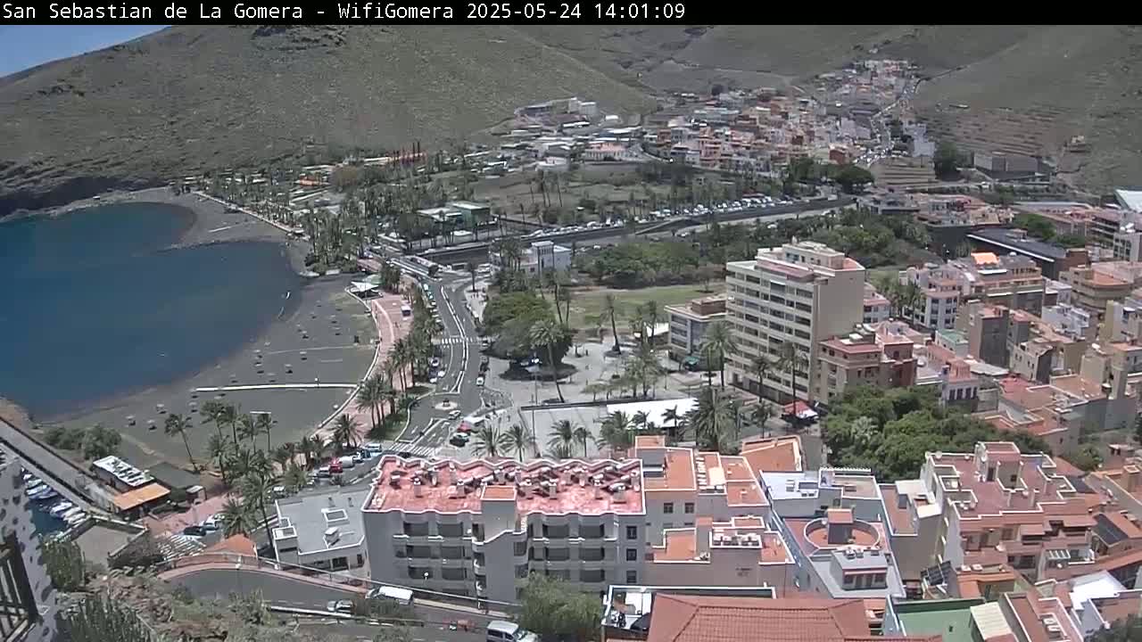 An aerial view shows a coastal town on a sunny day, with a dark-sand beach bordering a calm body of water and buildings extending up a hillside.
