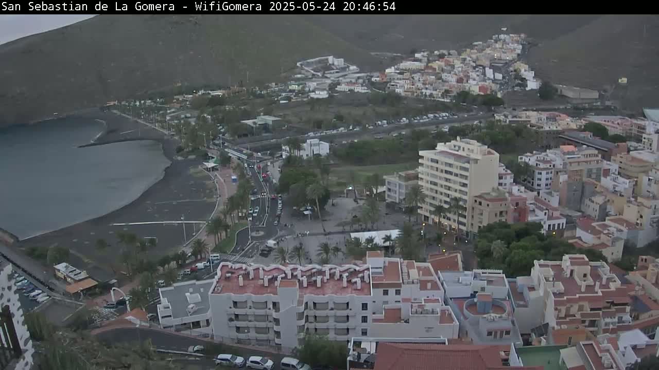 An aerial view shows a coastal town at dusk, with dark water bordering a black sand beach and numerous buildings rising up a hillside.