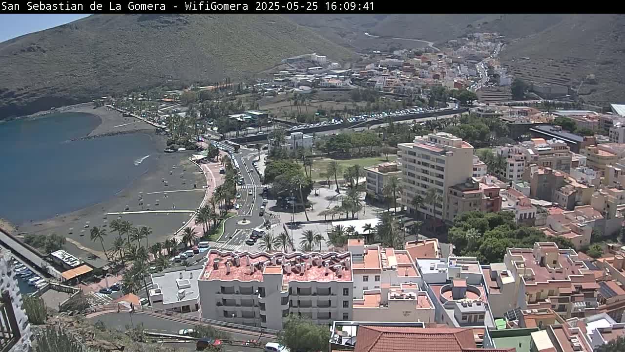An aerial view of a coastal town on a sunny day shows a dark-sand beach, a calm bay, palm trees, buildings, and roads.