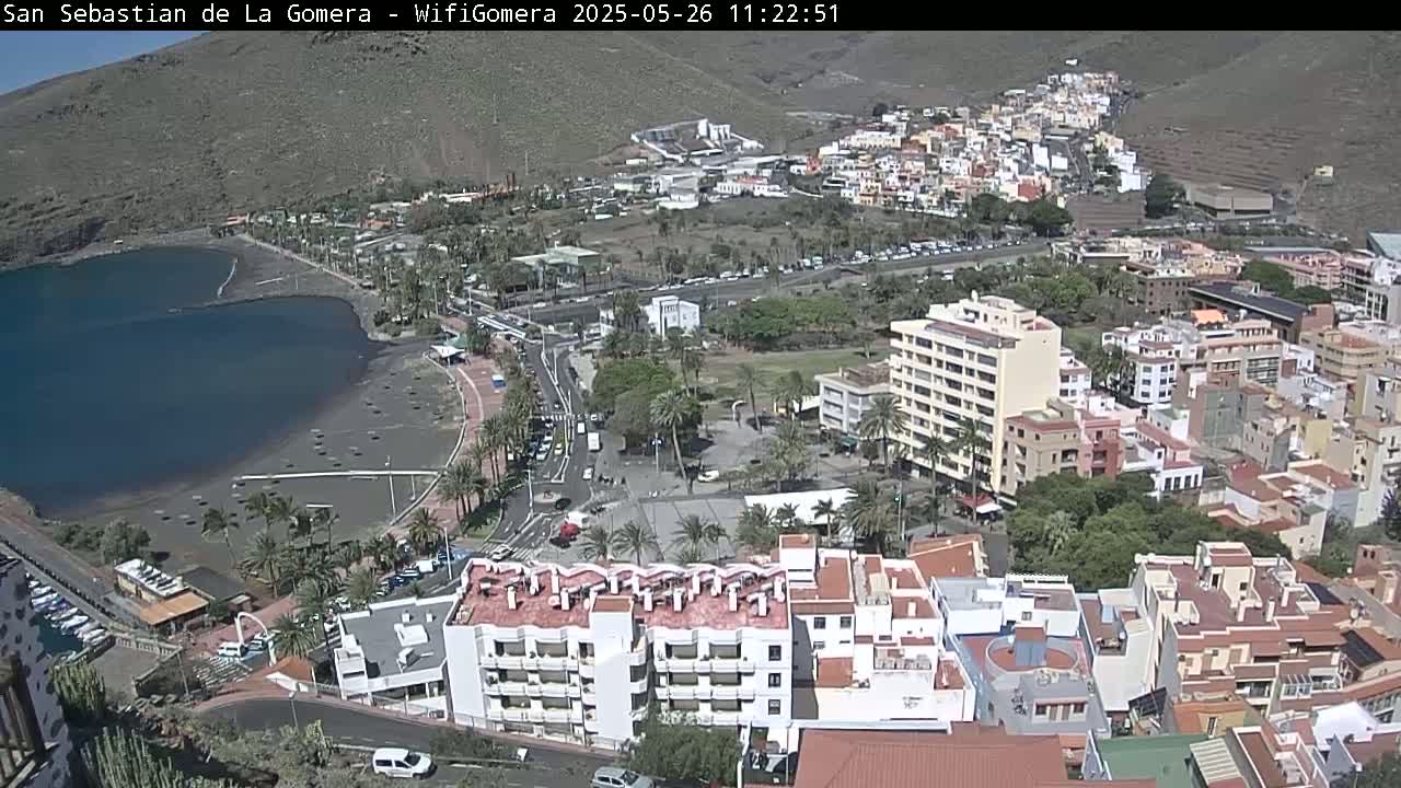 An aerial view of a coastal town on a sunny day, showing a dark-sand beach, a calm body of water, and numerous buildings nestled against a hillside.