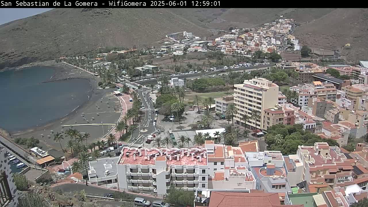 An aerial view shows a coastal town with a dark-sand beach and calm ocean under a mostly sunny sky, with buildings sprawling uphill from the shore.
