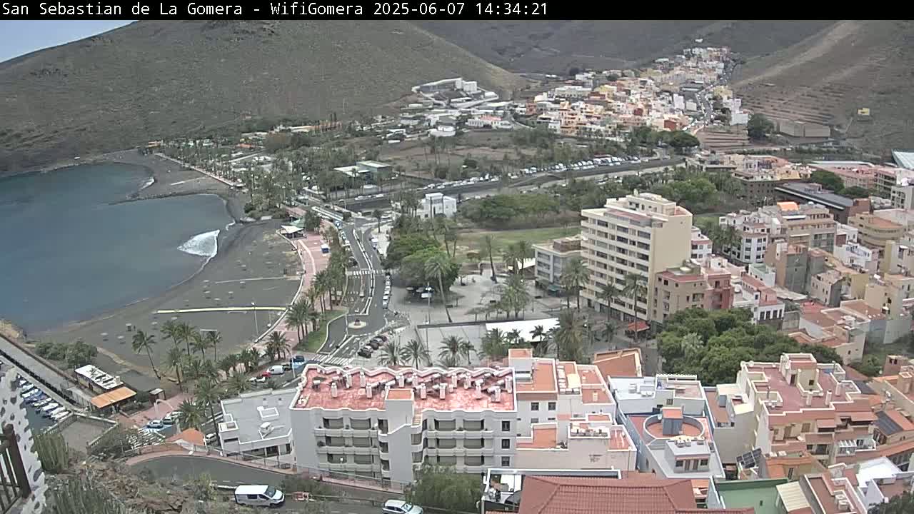 An aerial view shows a coastal town on a sunny day, with a dark-sand beach bordering a calm bay and buildings cascading up a hillside.