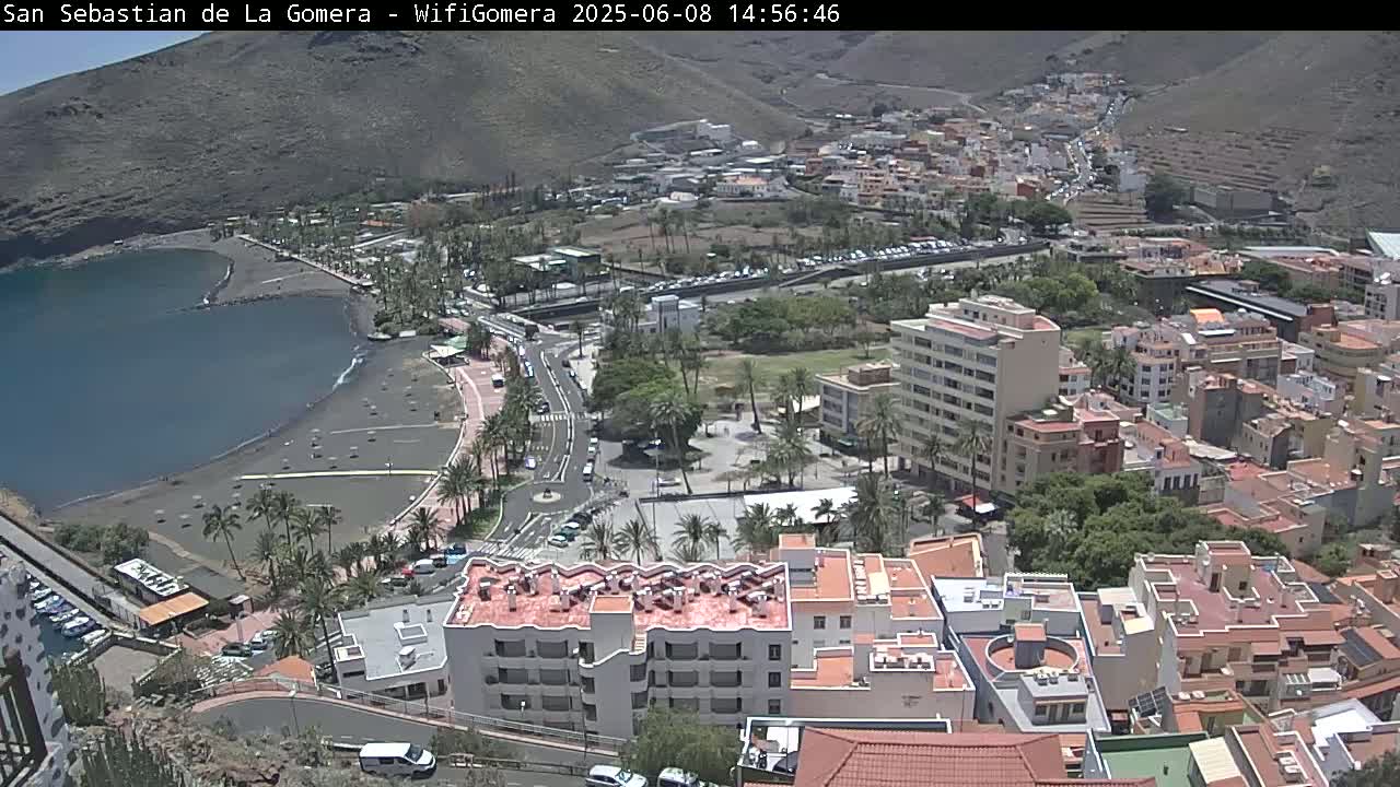 An aerial view shows a coastal town on a sunny day, with a dark-sand beach bordering a calm bay, and numerous buildings extending up a hillside.