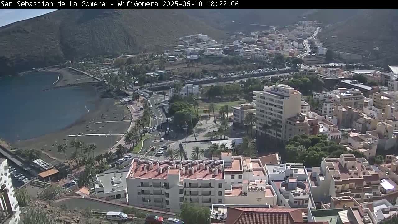 An aerial view shows a coastal town on a sunny day, featuring a dark-sand beach next to a calm body of water, and buildings extending up a hillside.