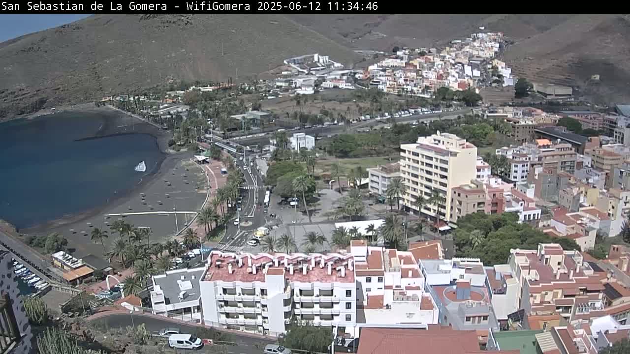 An aerial view shows a coastal town on a sunny day, with a dark-sand beach bordering a calm body of water, and numerous buildings stretching up a hillside.