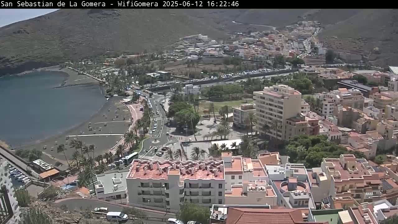 A coastal town with a dark-sand beach and a calm bay is seen from an elevated viewpoint on a sunny day.
