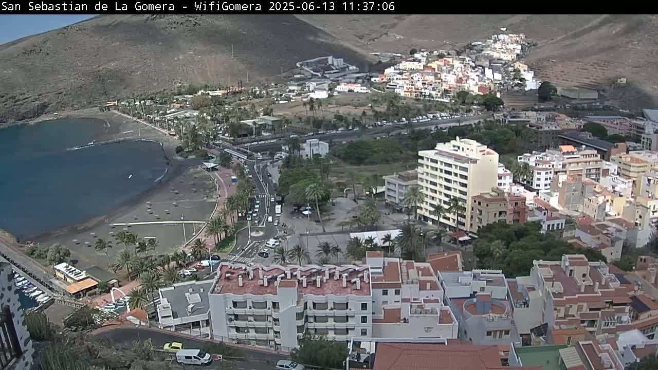 An aerial view shows a coastal town with a dark-sand beach, buildings clustered together, palm trees, and clear skies.