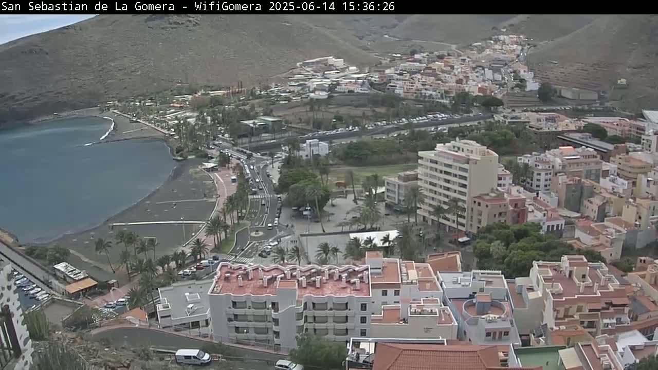 An aerial view shows a town situated on a hillside overlooking a dark-sand beach and calm ocean under an overcast sky.
