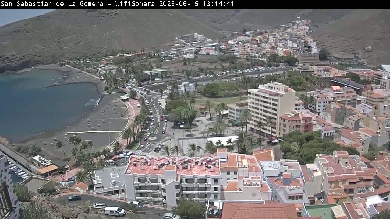 A coastal town with a dark-sand beach and calm ocean under a mostly sunny sky, nestled between hillsides.