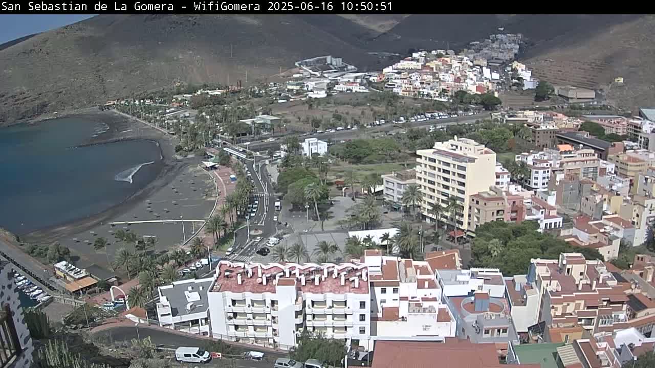 An aerial view shows a coastal town with a dark-sand beach, buildings, palm trees, and clear skies.