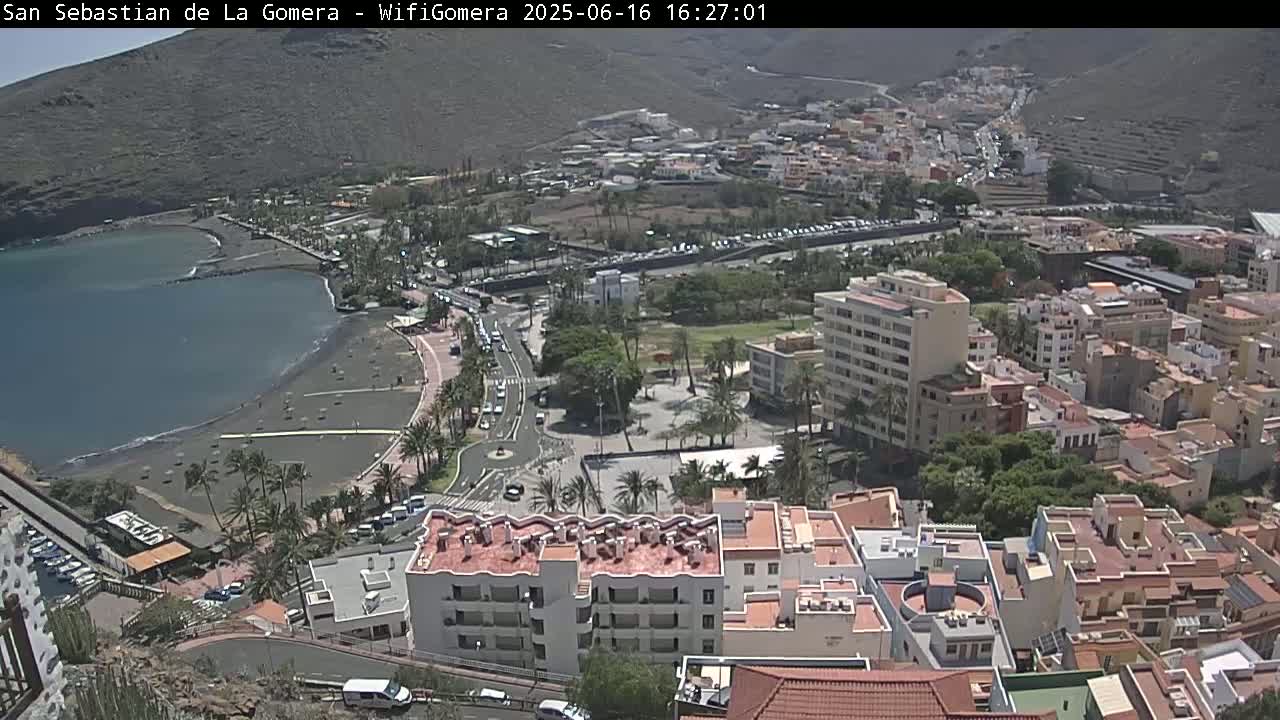 An aerial view shows a coastal town on a sunny day, with a dark-sand beach next to a calm body of water, and numerous buildings stretching up a hillside.