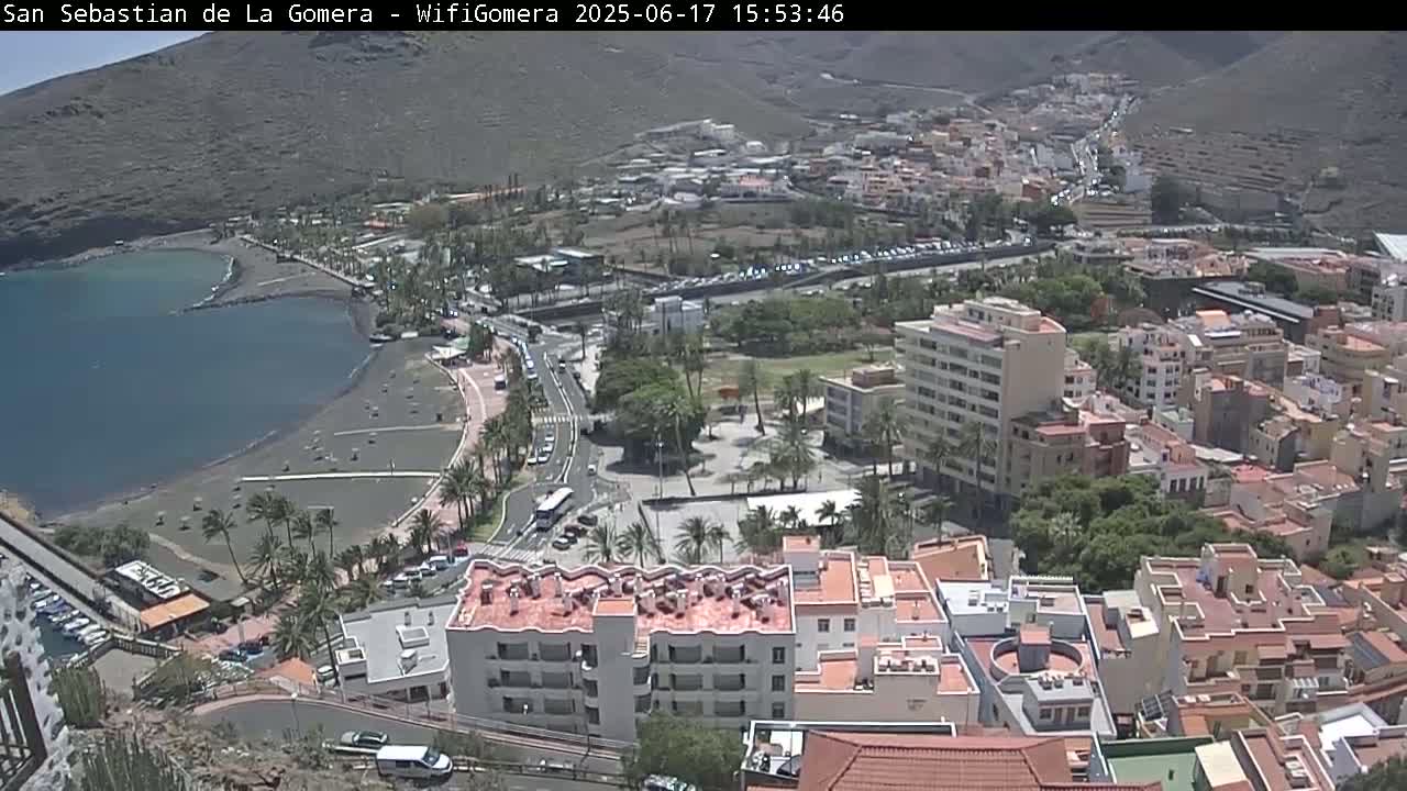 An aerial view shows a coastal town with a dark-sand beach, buildings, palm trees, and clear sunny skies.
