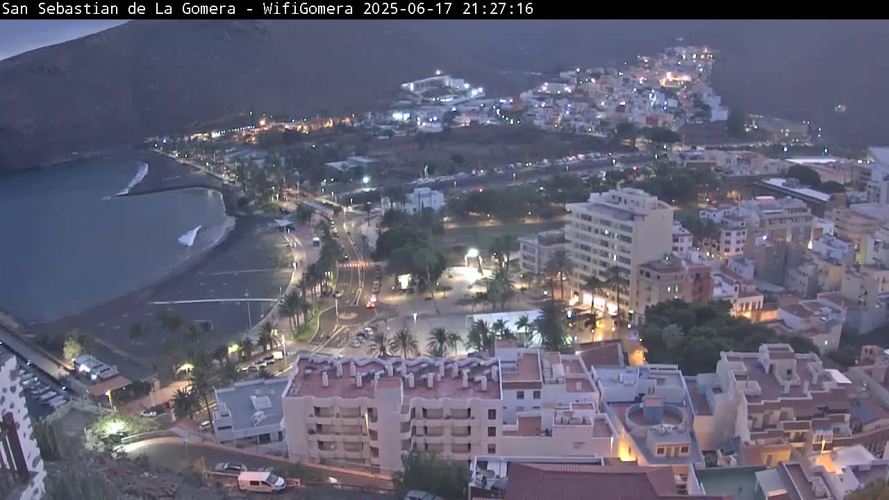 A nighttime view of a coastal town, showing a dark-sand beach, calm ocean waters, and buildings illuminated against a dusky sky.