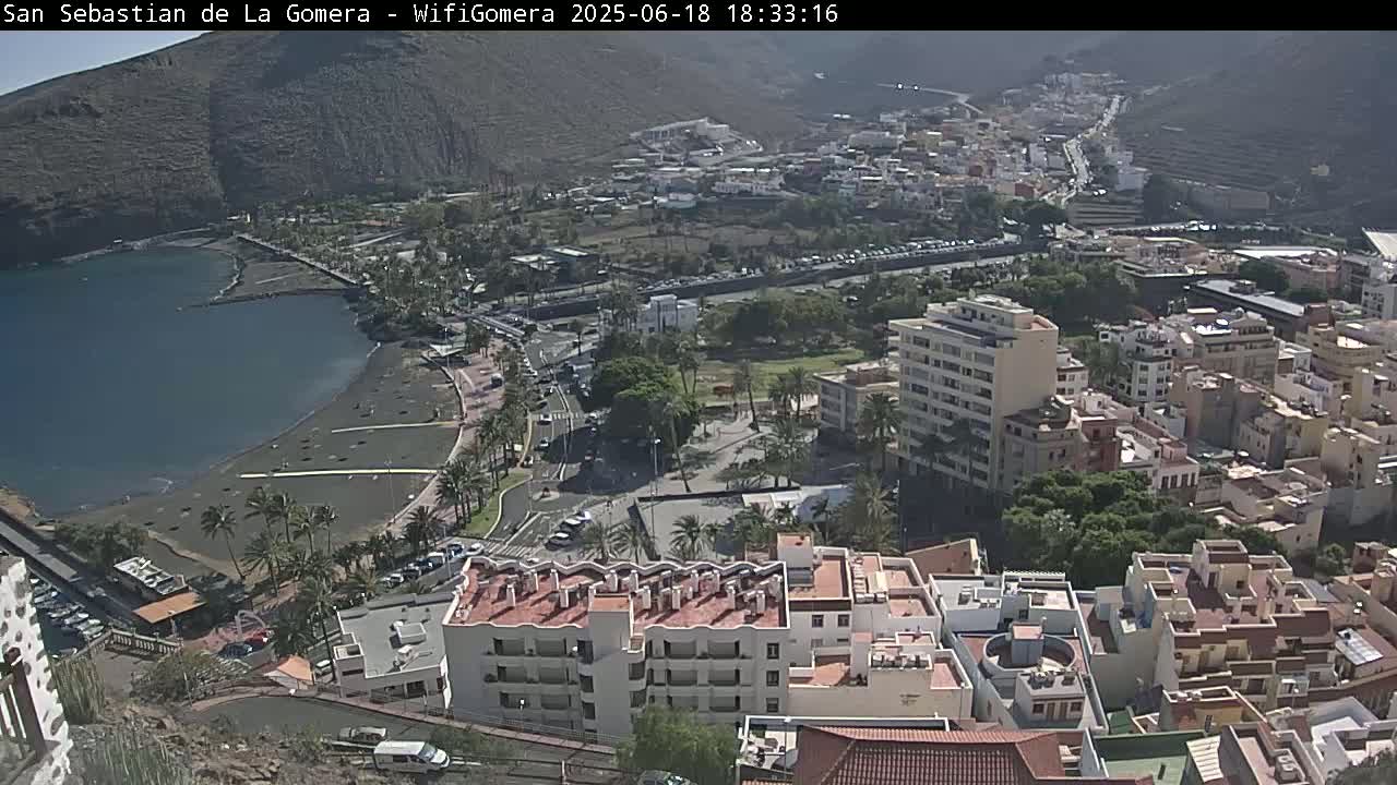 A coastal town with a black sand beach, palm trees, and multi-story buildings is visible under sunny conditions.