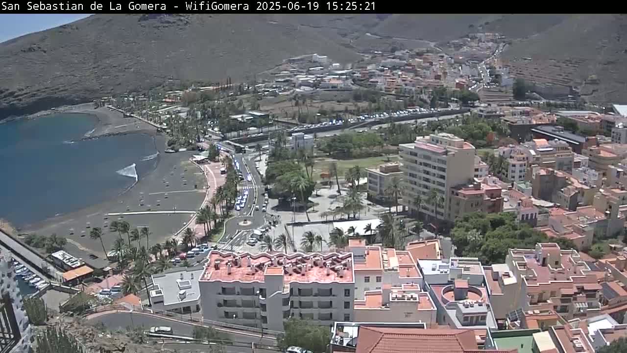 An aerial view shows a coastal town on a sunny day, with a dark-sand beach beside calm ocean water and buildings extending uphill.