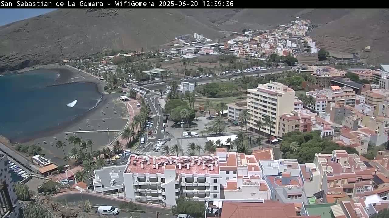 An aerial view shows a coastal town on a sunny day, with a dark-sand beach, calm ocean water, and numerous buildings extending up a hillside.