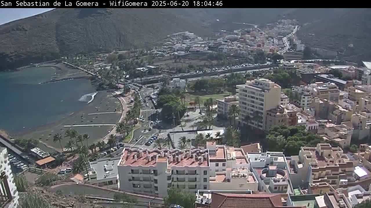 An aerial view shows a coastal town on a sunny day, with a dark-sand beach and calm ocean water bordering a collection of buildings and palm trees.