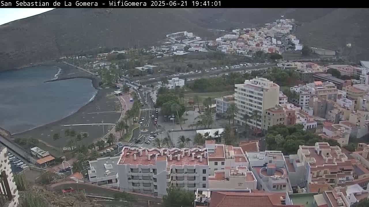 An overcast day reveals a coastal town with a dark-sand beach and calm ocean waters, nestled among buildings and hillsides.