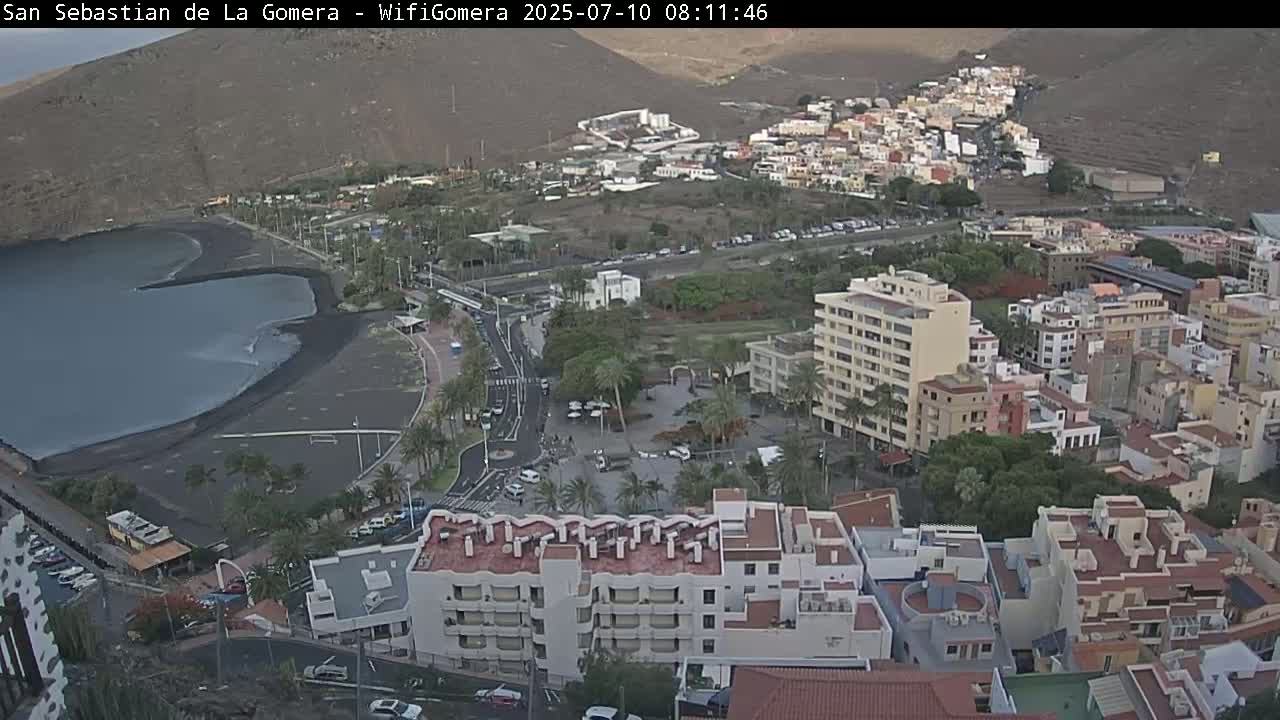 An aerial view shows a coastal town with a dark-sand beach and calm ocean, under mostly clear skies.