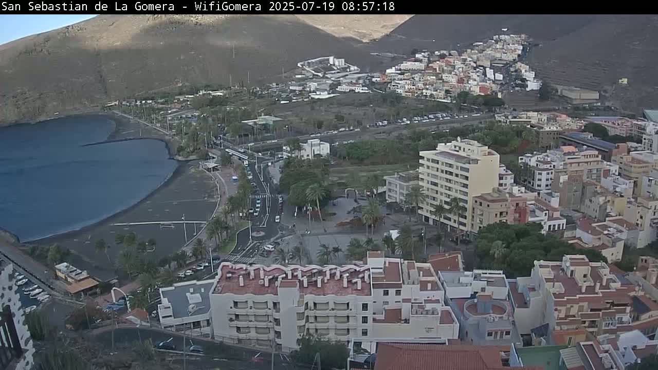 An aerial view shows a coastal town with dark-blue water, a black sand beach, buildings, palm trees, and parking lots under mostly clear skies.