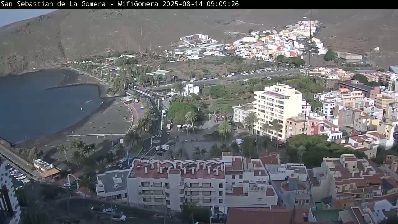 An elevated view shows a coastal town with a dark-sand beach and calm water, under a mostly clear sky.